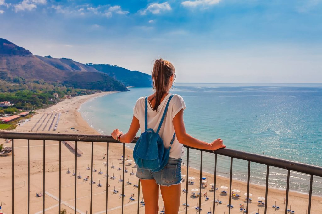 Donna con zaino azzurro osserva il mare da un balcone panoramico sulla spiaggia.