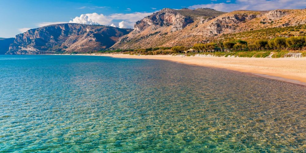 Spiaggia sabbiosa con acque cristalline e monti sullo sfondo.